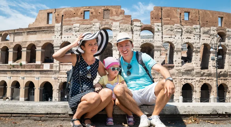Parents and a toddler in a sun hat sitting on a wall in front of the Colosseum