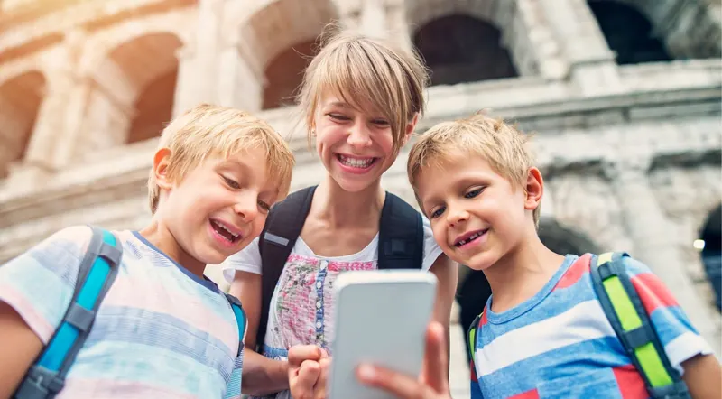 Three happy kids with backpacks looking at a phone outside the Colosseum